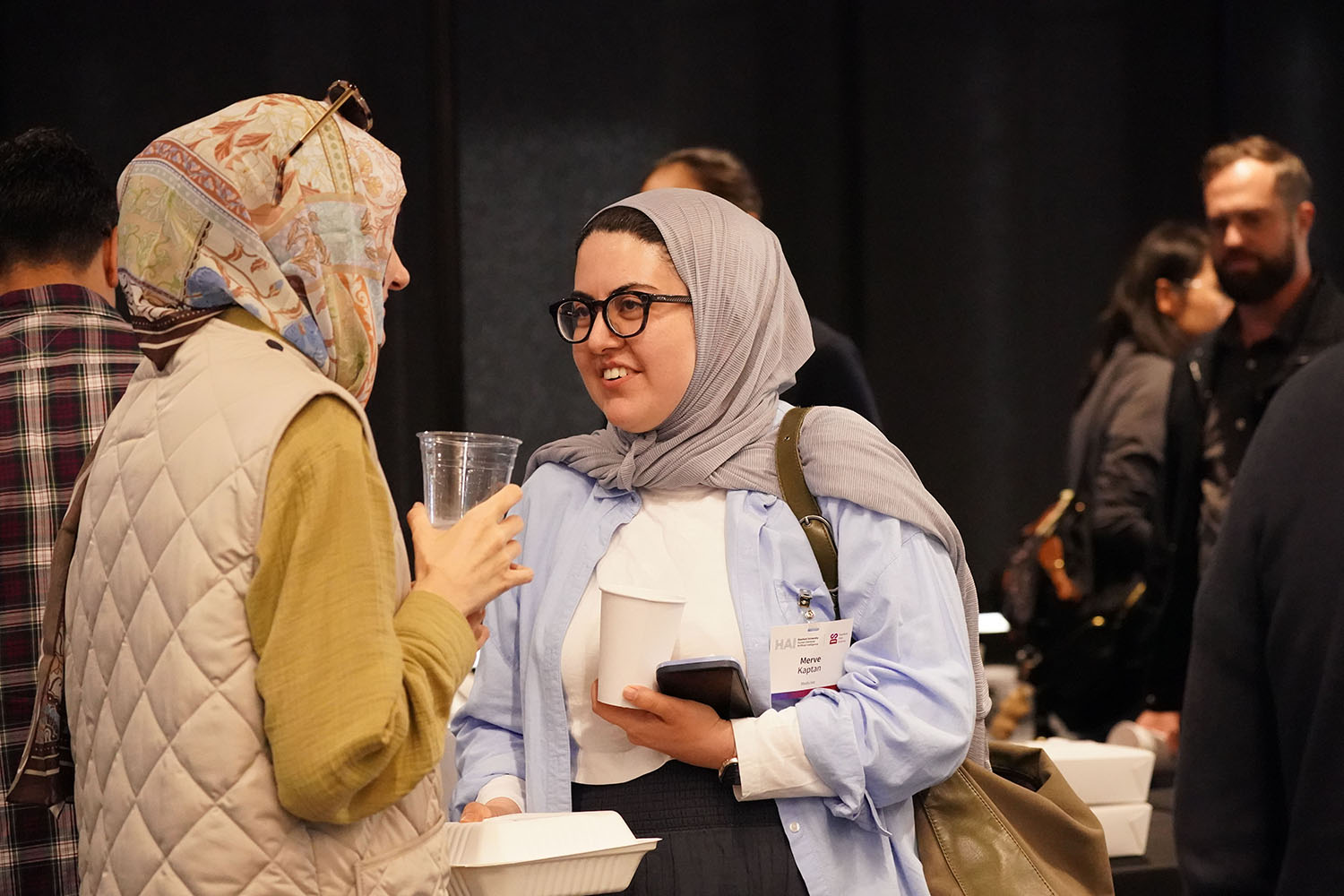 Two women in headscarves talk at CORES symposium, one holding a cup and the other a food container and cup, in a busy background.