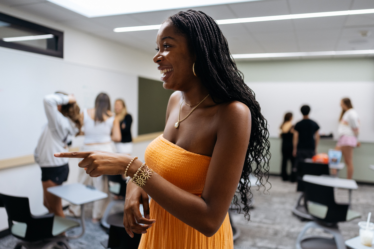 Gabby Edelin gestures while standing in a classroom, with other students in the background engaging with each other.