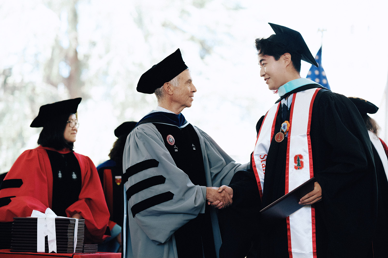 A graduate in cap and gown shakes hands with the dean during a graduation ceremony on stage.