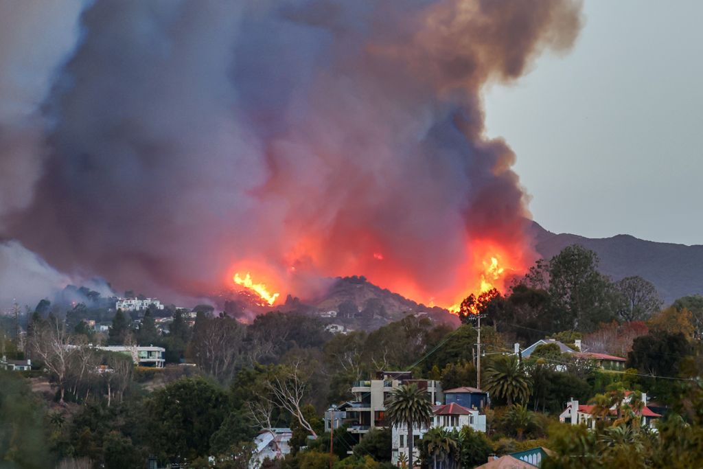 Wildfire smoke billows above hills, with flames visible through trees near residential areas.