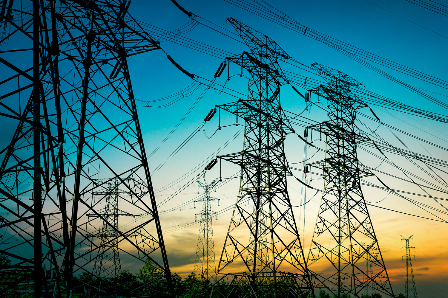 High-voltage power lines stretch across the sky during sunset, silhouetted against a vibrant blue and orange backdrop.