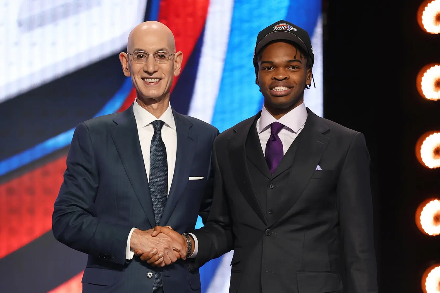 NBA commissioner Adam Silver shaking hands Ebuka Okorie, both of them wearing formal attire and smiling into the camera.