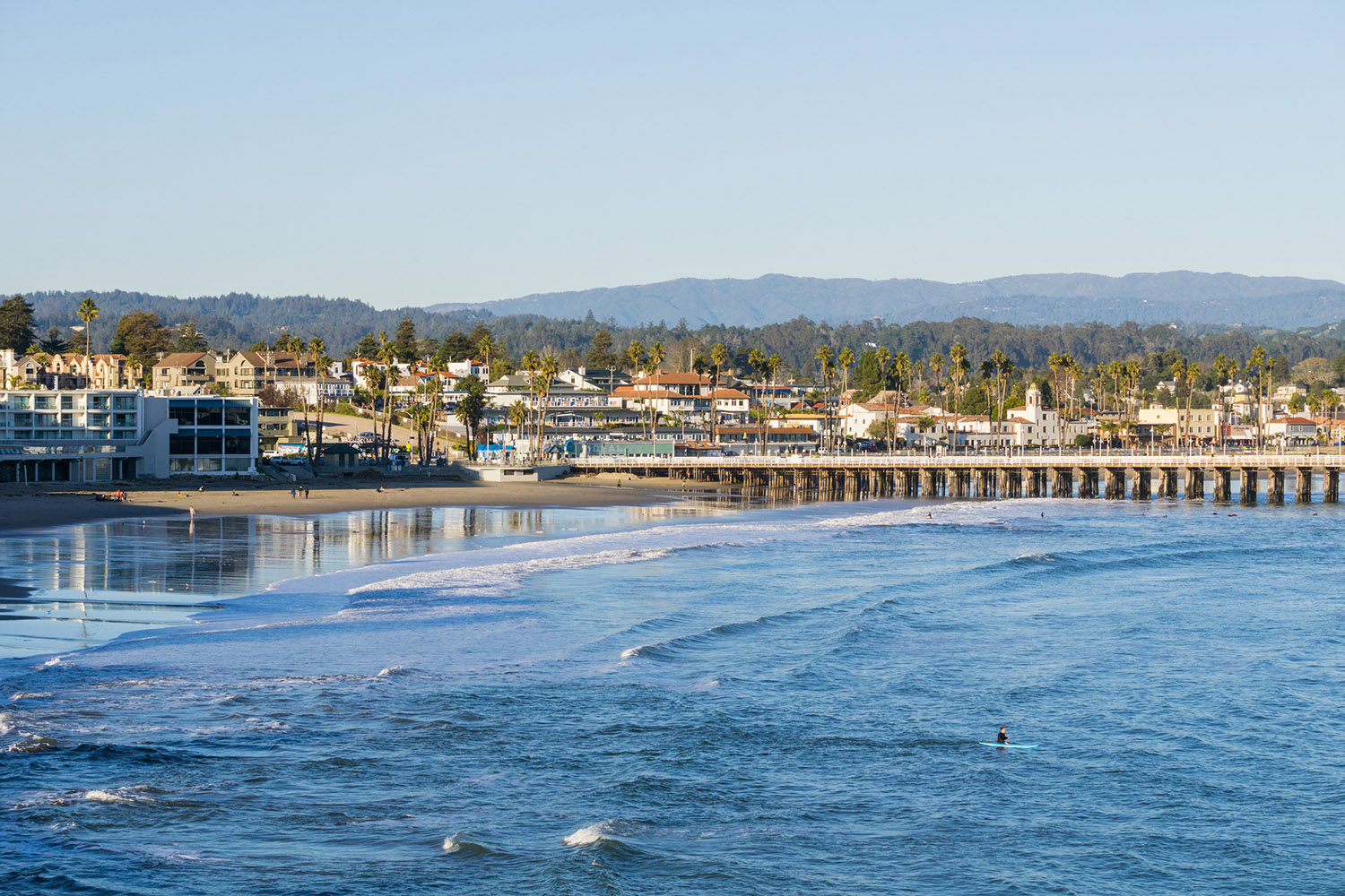 Coastal scene of Santa Cruz, CA, featuring a beach, pier, and palm trees under a blue sky, with a surfer in the water.