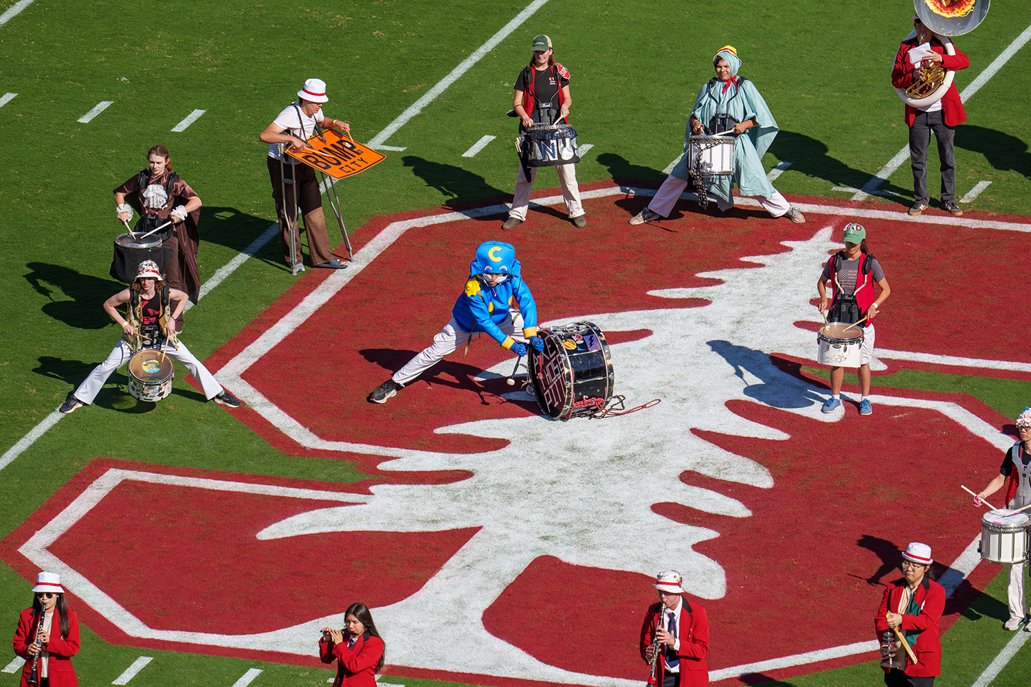 Marching band members in costume play drums and instruments while forming a shape on the football field.