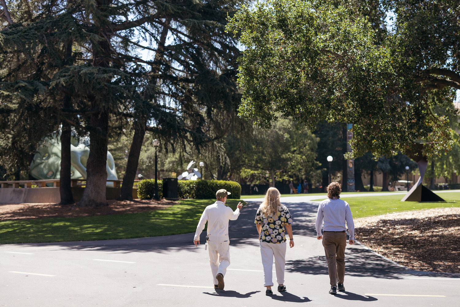 Art ranger Mark Shunney leads two visitors down a campus path past outdoor sculptures on a sunny day.