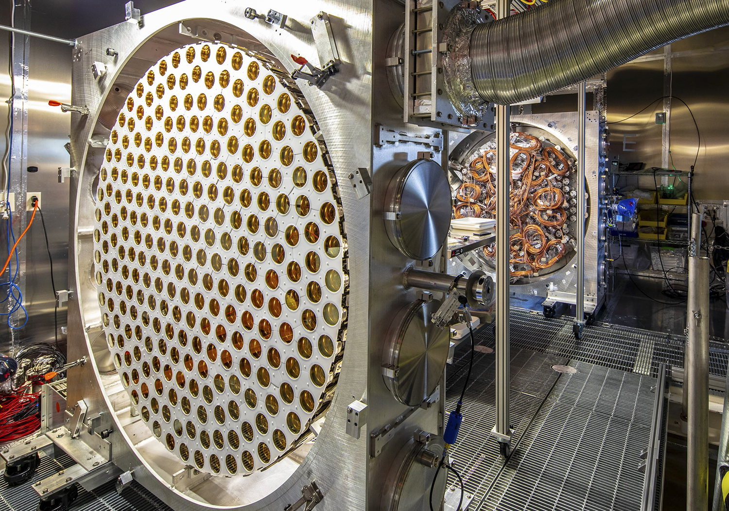 A large circular detector panel studded with gold sensors stands open in a metal-walled laboratory room.