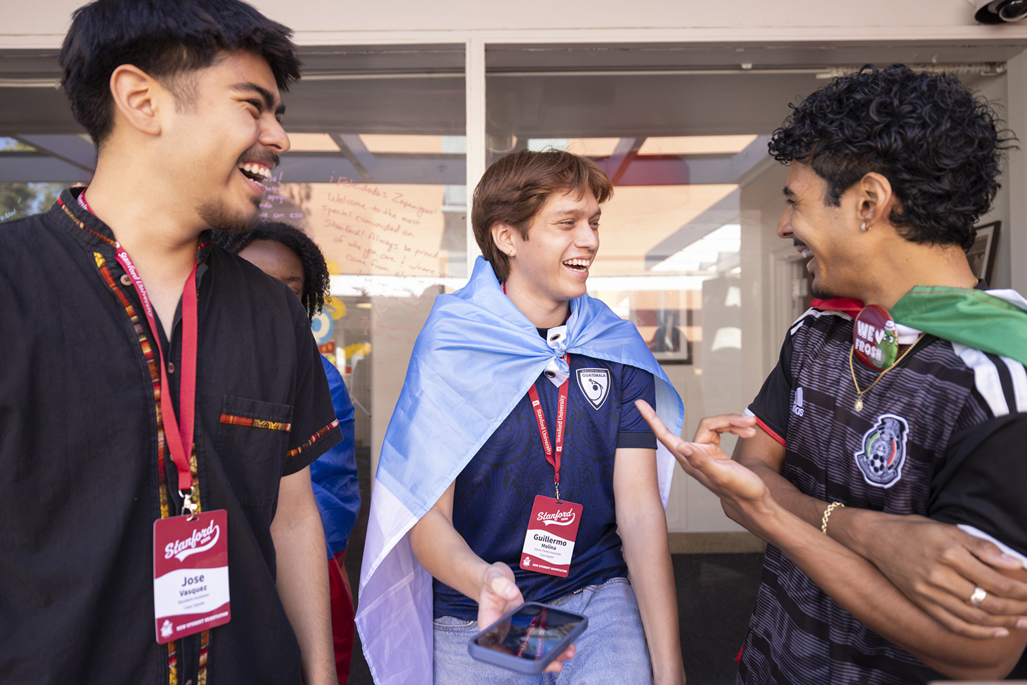 Three students wrapped in flags from their home countries laugh together during a welcome gathering.