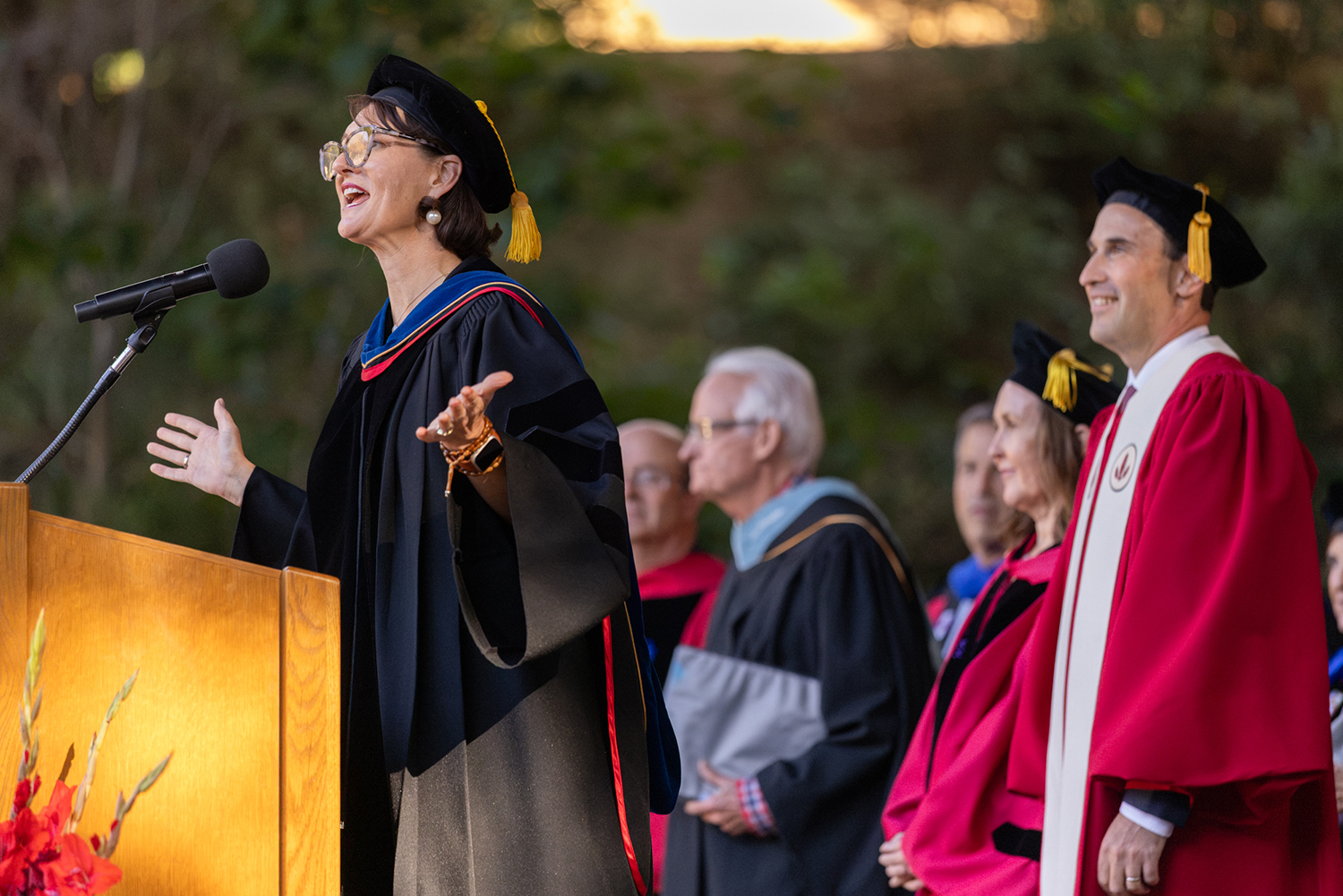 Rev. Dr. Tiffany Steinwert in academic robes speaks warmly at a podium as colleagues look on.