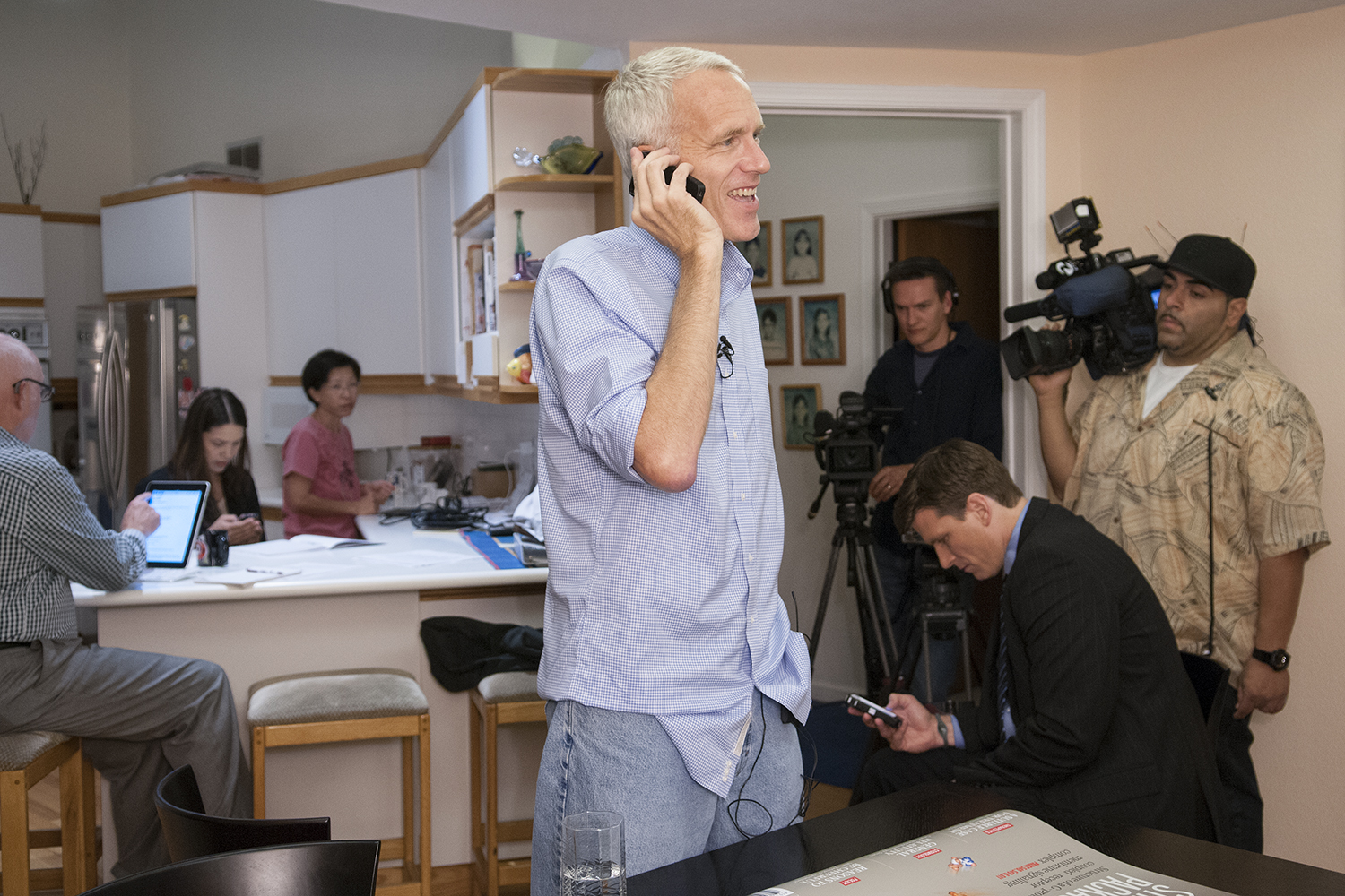 Brian Kobilka on a phone call stands in a kitchen as camera crews and reporters wait nearby.