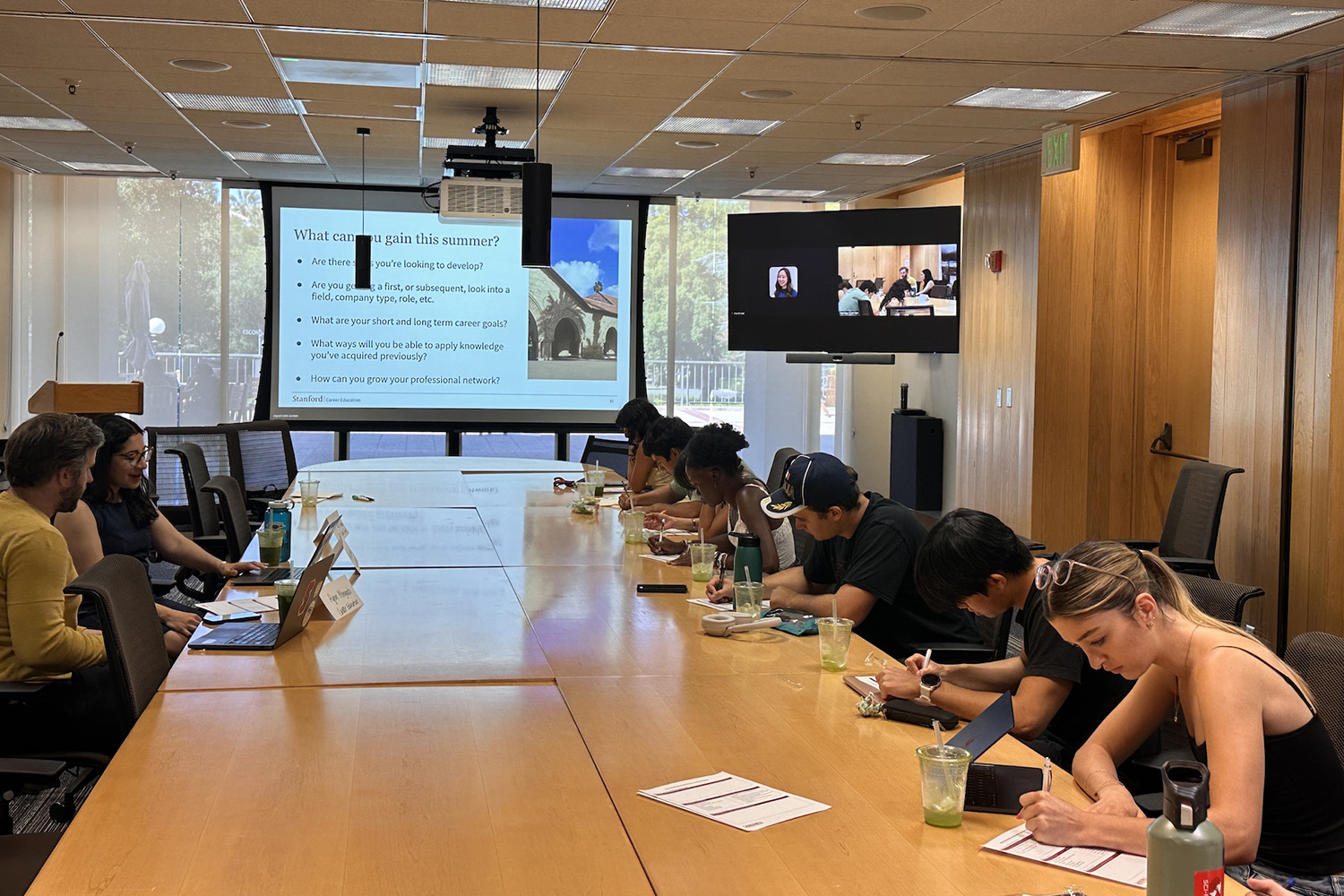 Students take notes at a long conference table during a career workshop, with slides and a video call displayed.