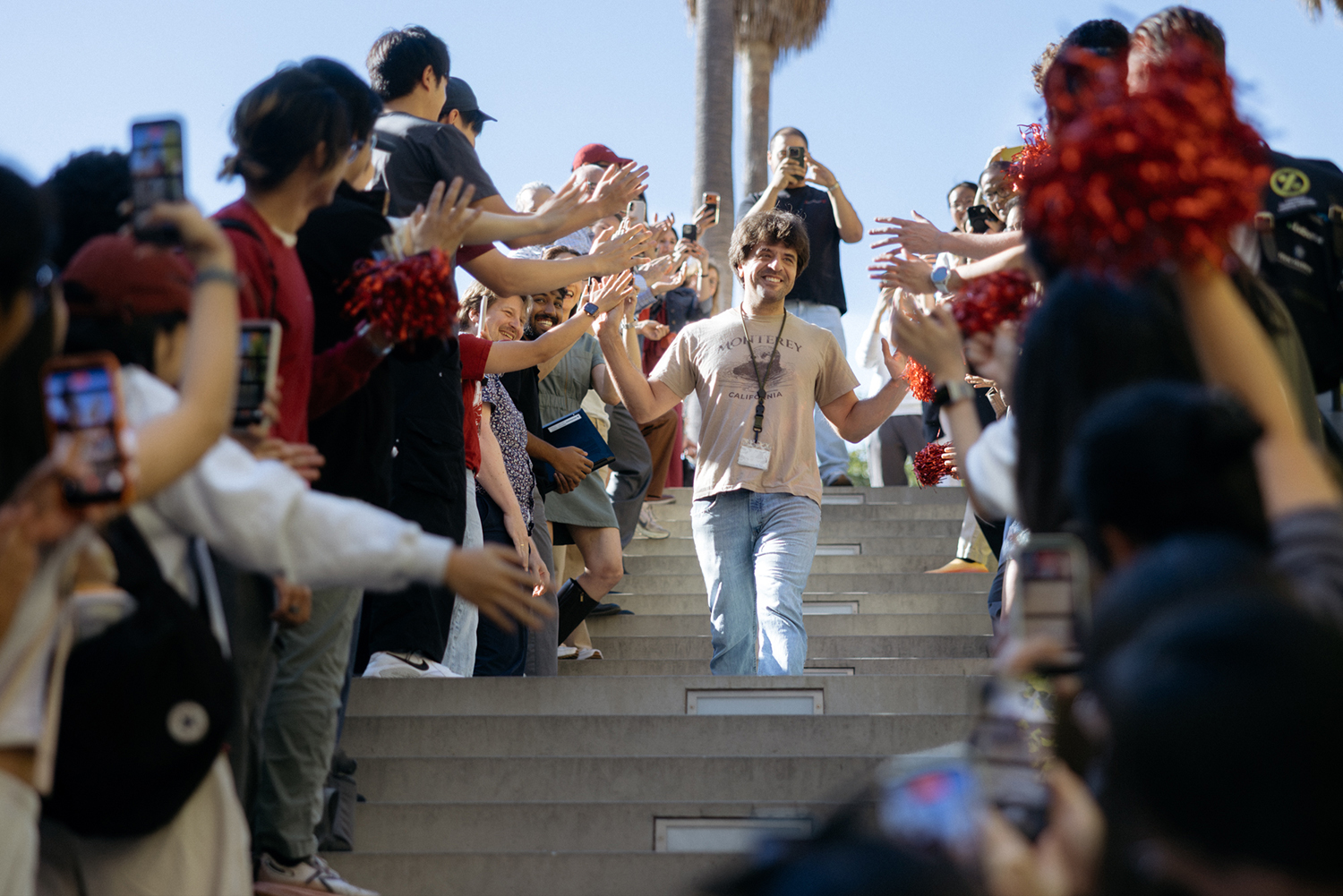 Karl Deisseroth walks down outdoor steps between two lines of cheering people holding red pompoms and phones.