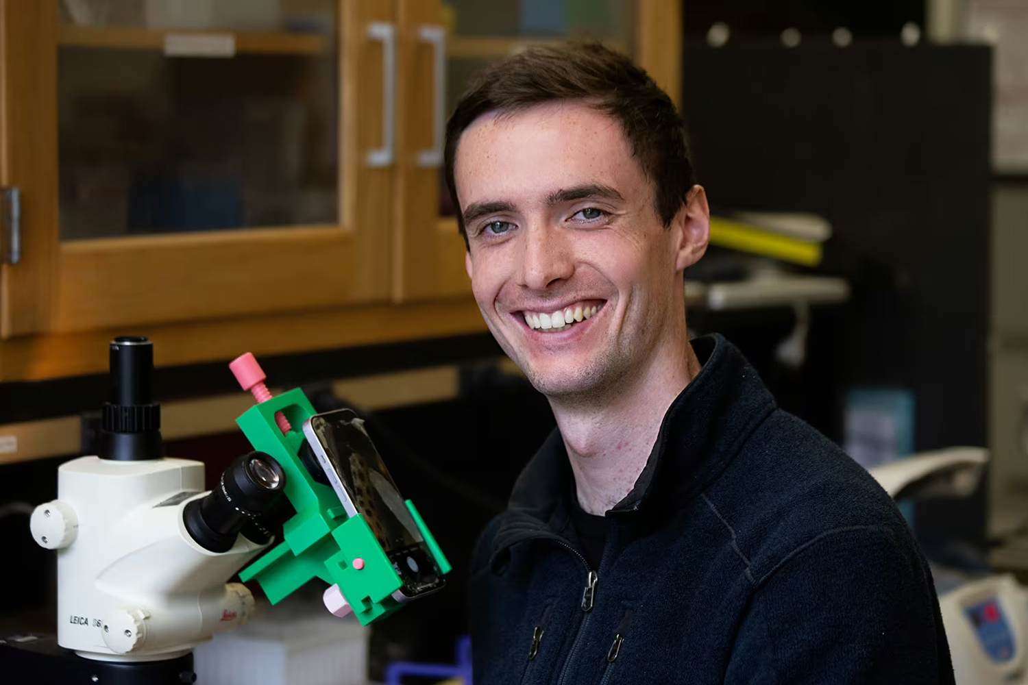Danny Collins sits beside a microscope fitted with a green 3D-printed mount that holds a smartphone.