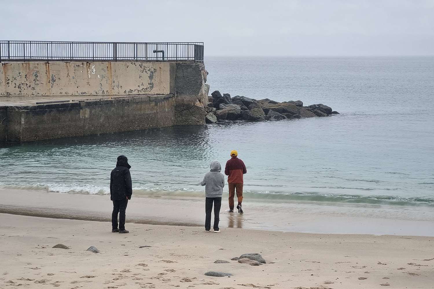 Three student in jackets stand at the water’s edge of a beach in Monterey Bay beside an old concrete seawall.