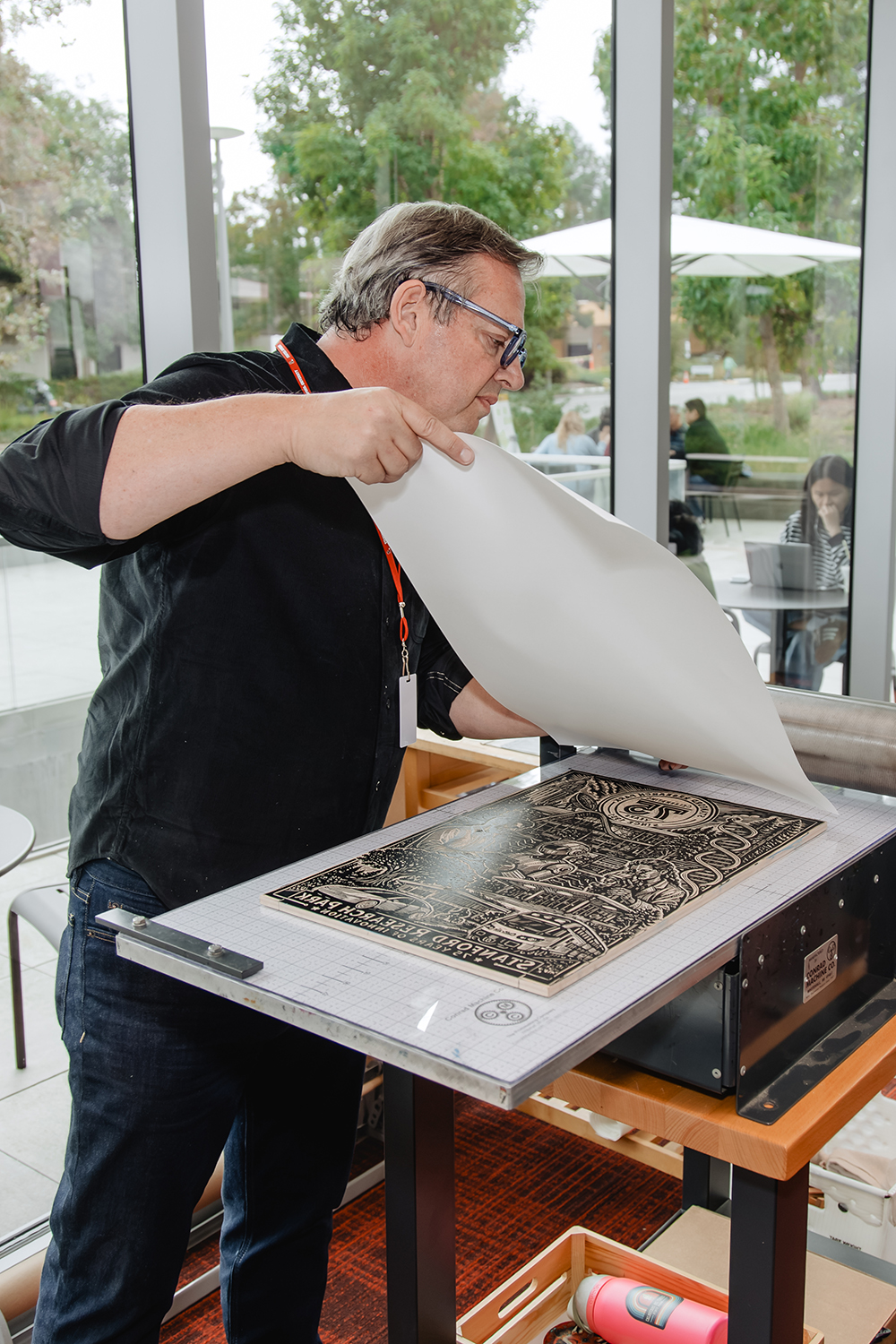 A printmaker lifts paper from an inked carved block bearing the Stanford Research Park 75th-anniversary design.