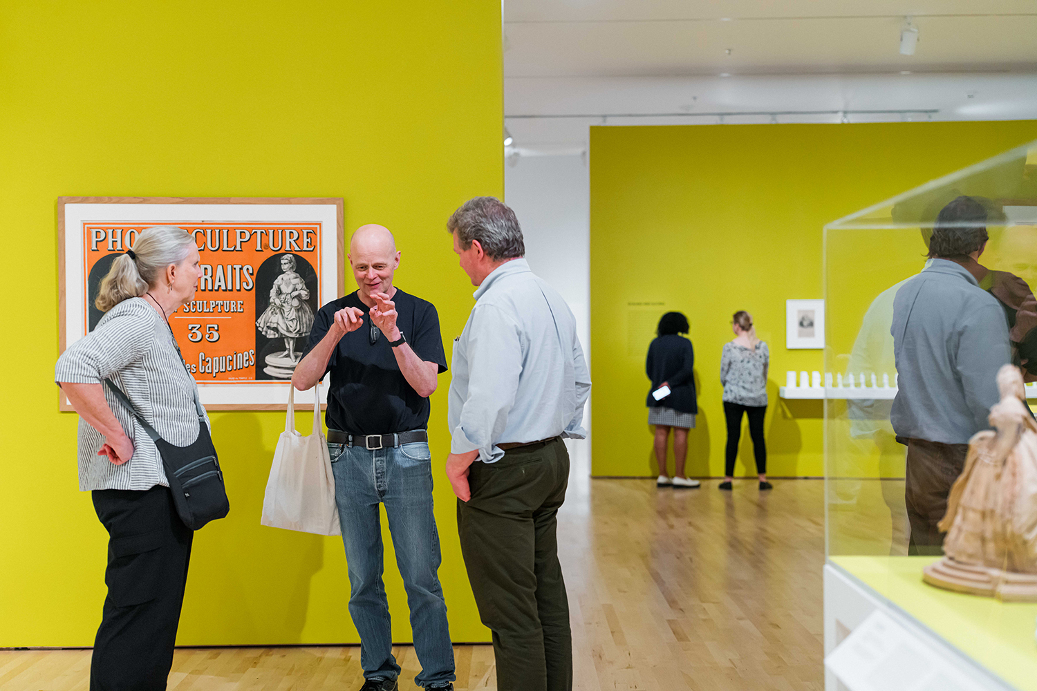 Three visitors talk in a gallery beside a framed vintage poster, with another display case and visitors visible beyond.