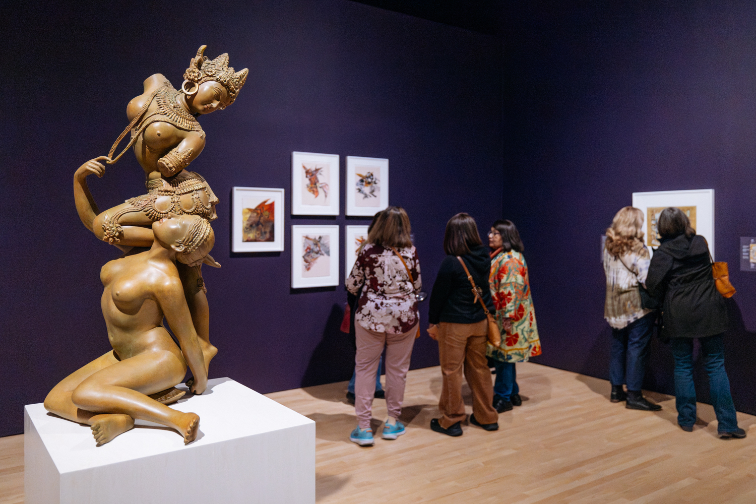 Bronze female figures are displayed on a white pedestal. Several visitors are admiring artworks in the background on a purple wall.