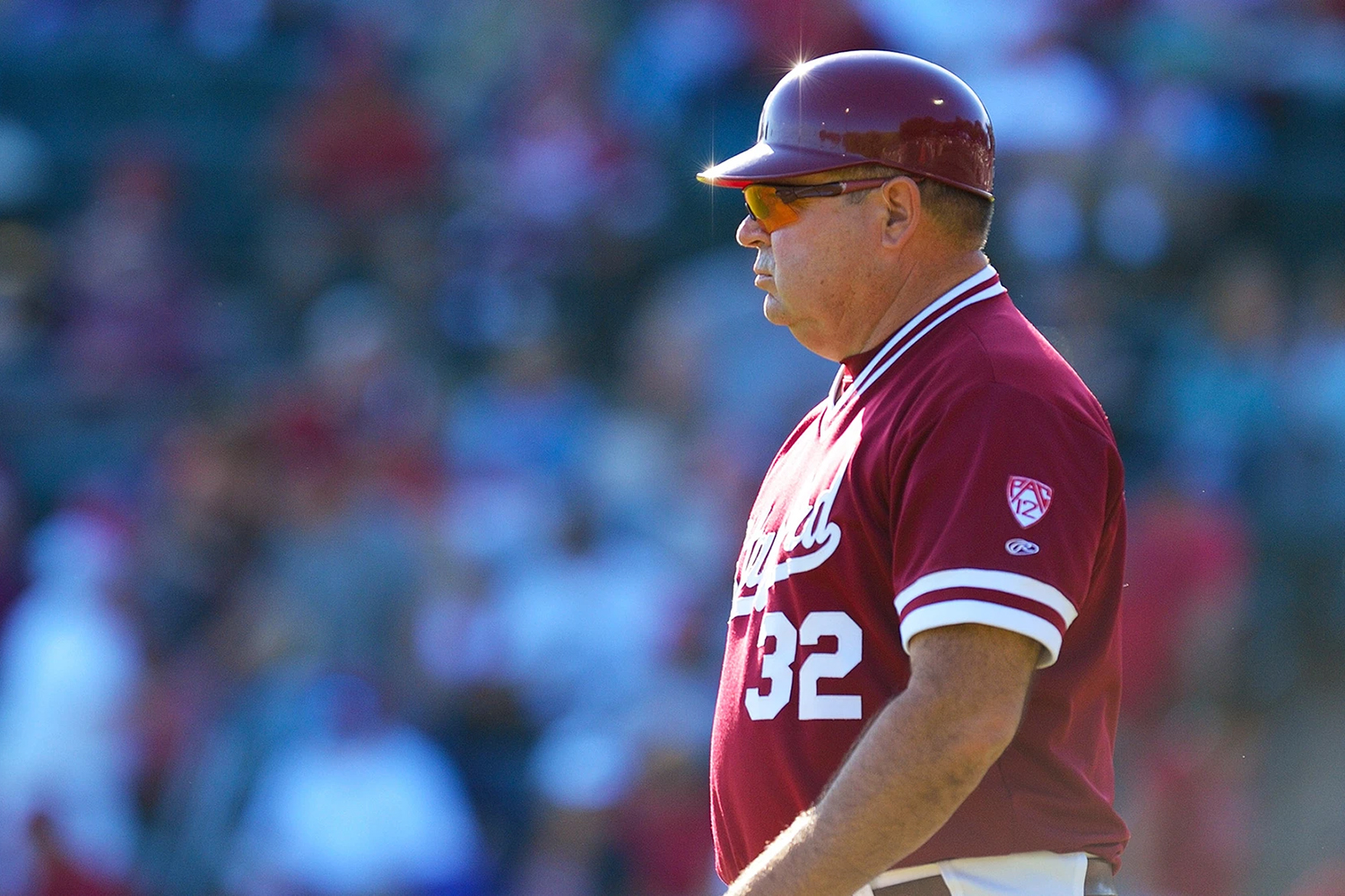 Baseball coach Dean Stotz in a maroon jersey observes the game under bright sunlight while fans are visible in the background.