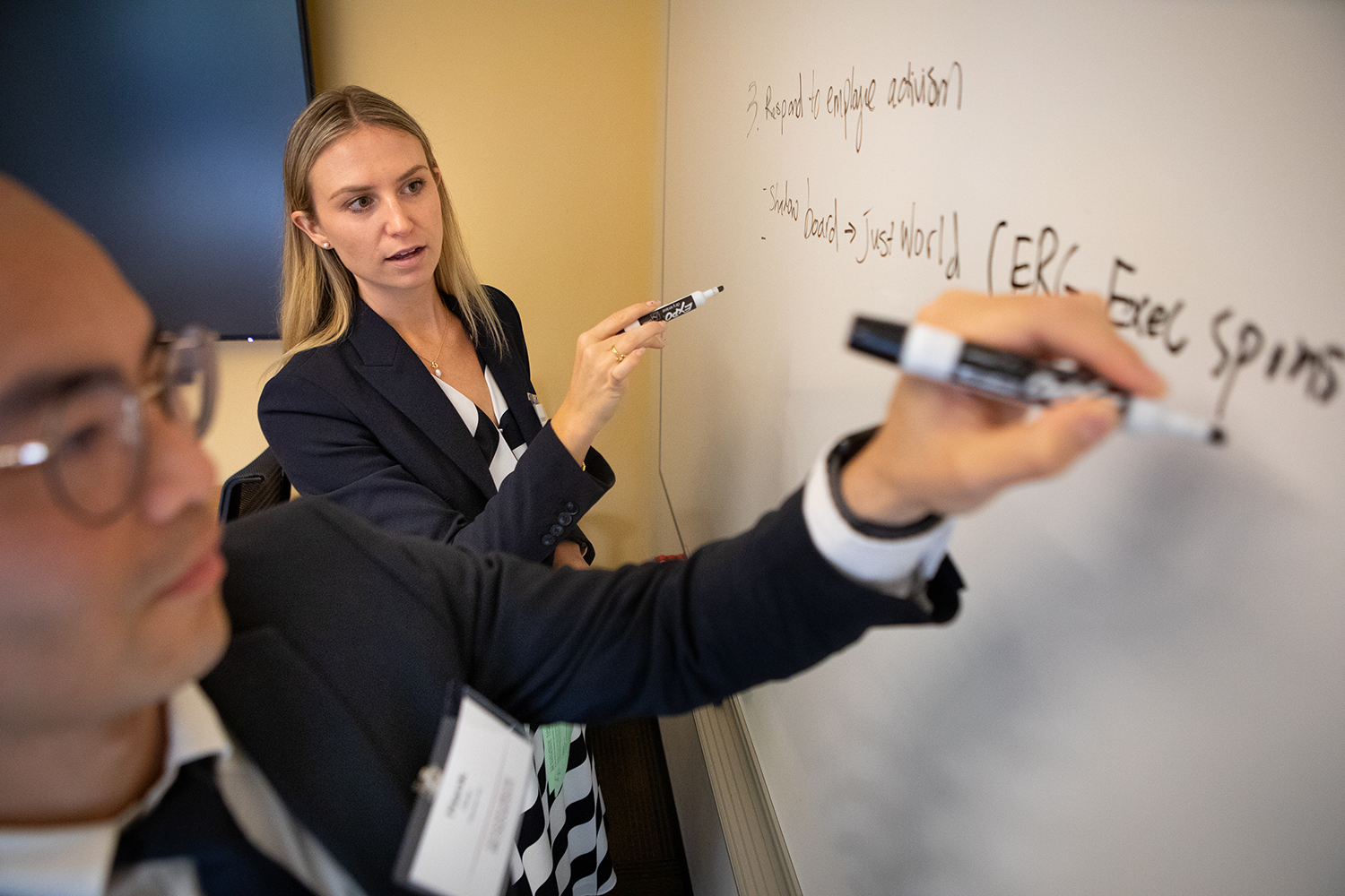 Two individuals writing on a whiteboard during a brainstorming session, focused on collaborative ideas and strategies in a meeting environment.