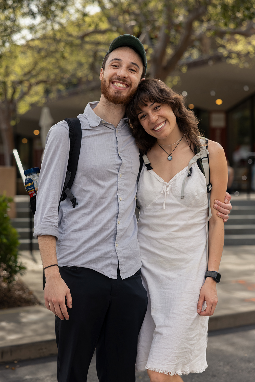Siblings stand together outdoors, smiling, with one wearing a button-up shirt and the other dressed in a casual summer dress.