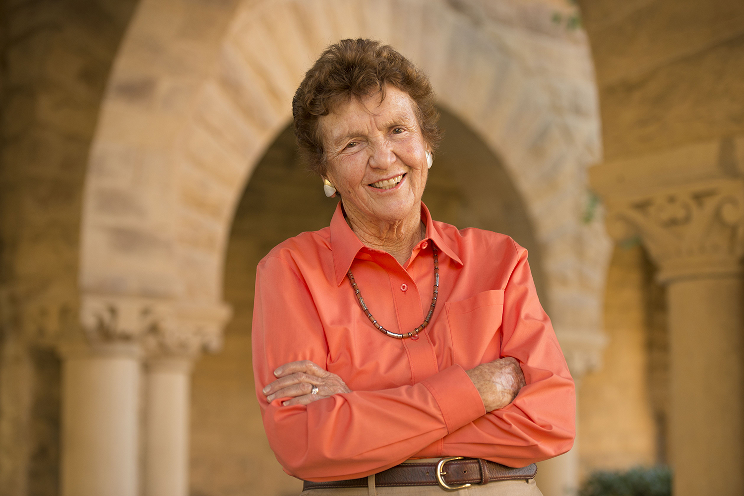 Joan Fletcher Lane standing with her arms crossed and smiling, wearing an orange blouse and necklace in a stone architectural setting.
