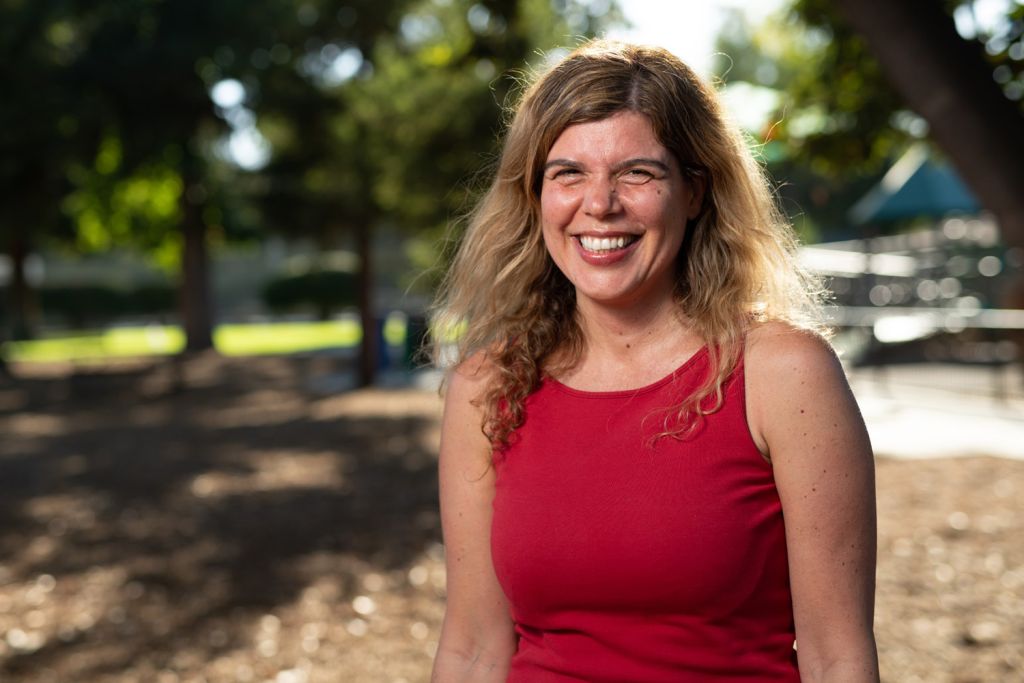 Anne Brunet with shoulder-length wavy blonde hair smiling outdoors in a red sleeveless top.