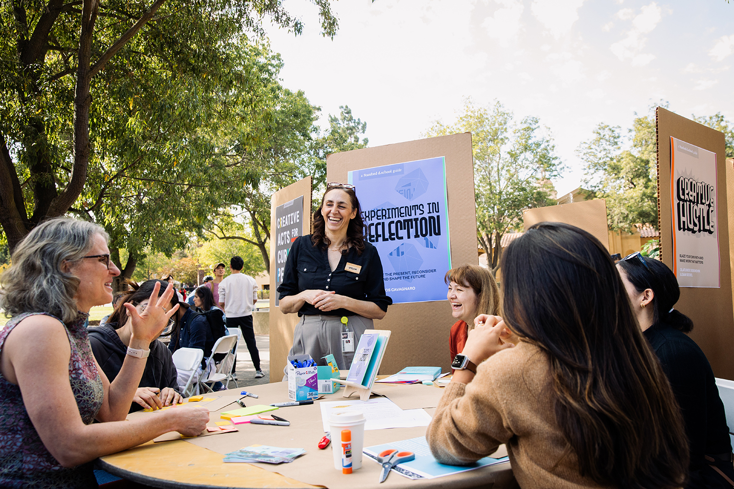 A woman leads a discussion at a gathering, with participants engaged around a table filled with sticky notes and materials for creative reflection.