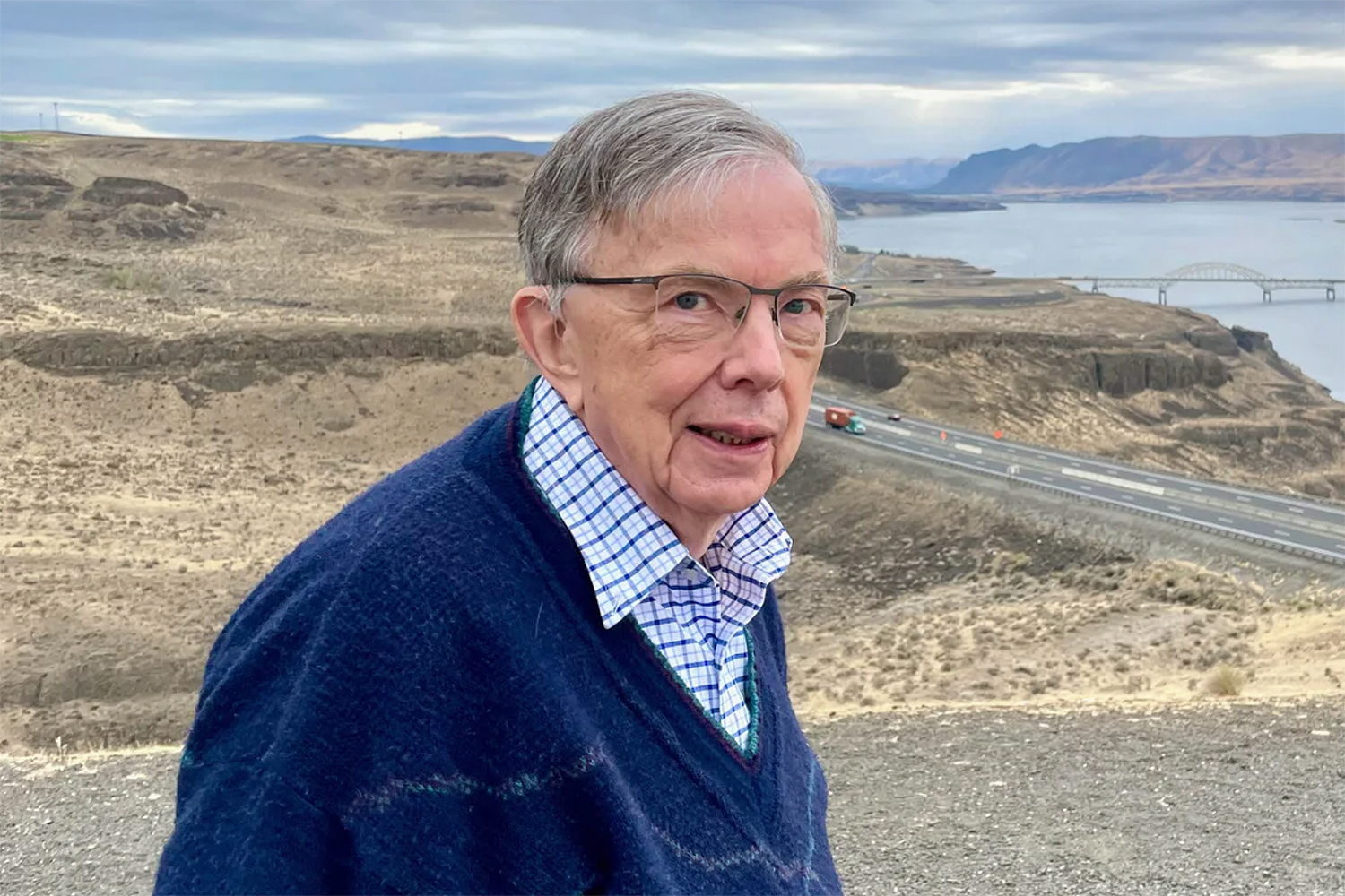 Frederick S. Hillier wearing a blue sweater stands near a scenic overlook, with a highway and river visible in the background.