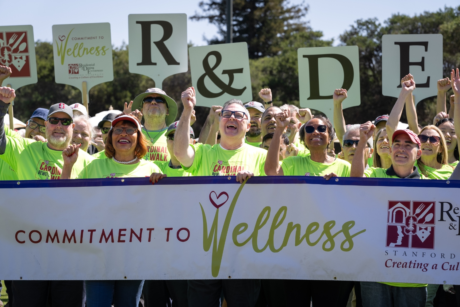 A group of people in bright green shirts holding signs and a banner promoting commitment to wellness at a community event.