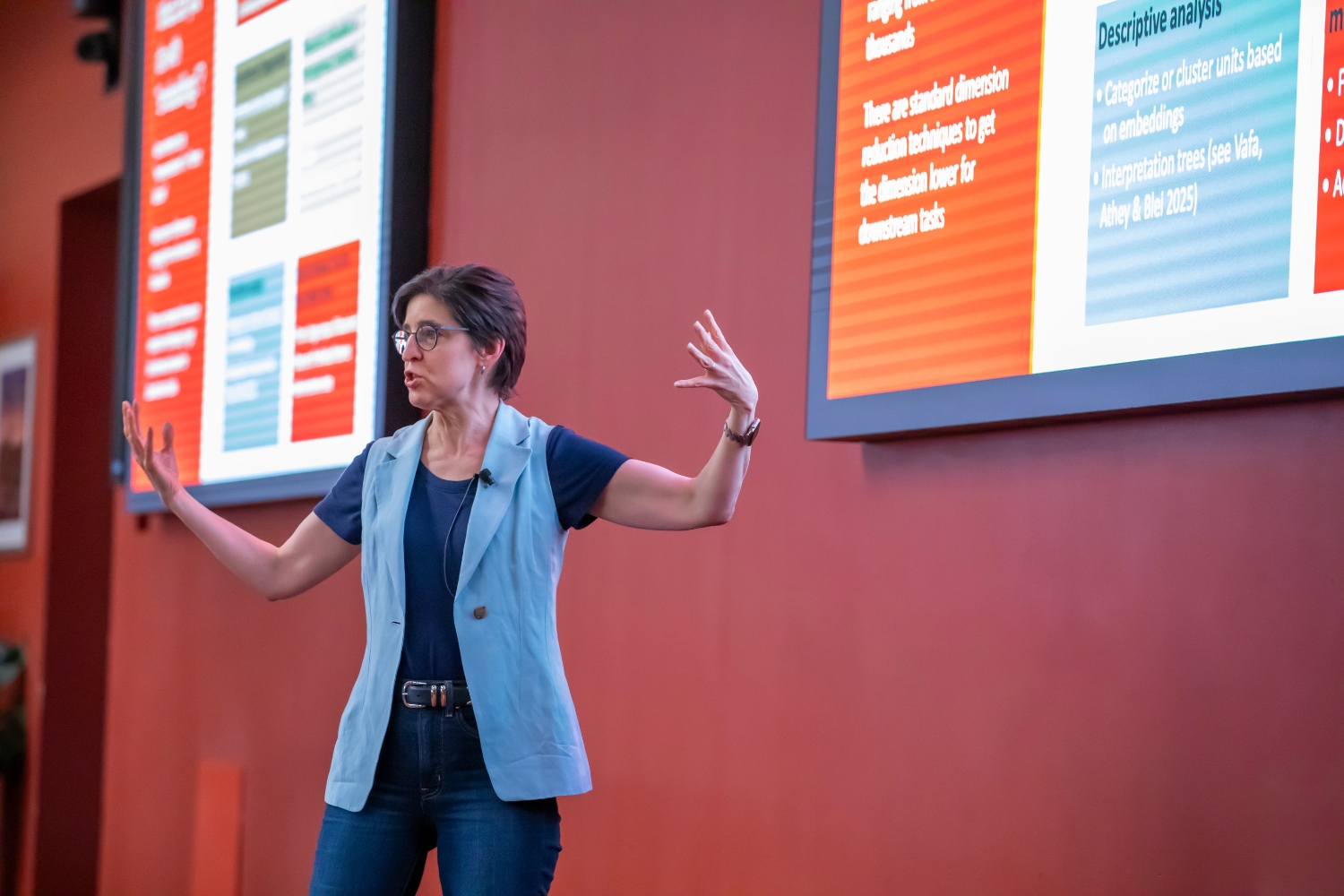 Susan Athey gestures while discussing the impact of AI on her work, with colorful slides displayed in the background.