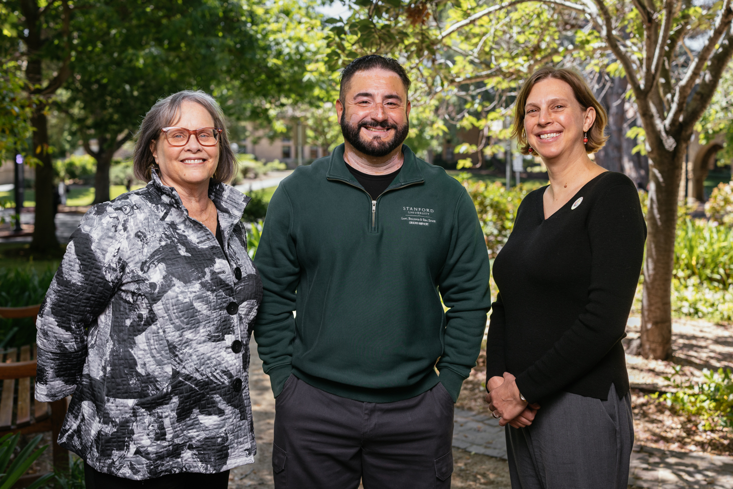 Three people smiling and standing together outdoors, surrounded by greenery.