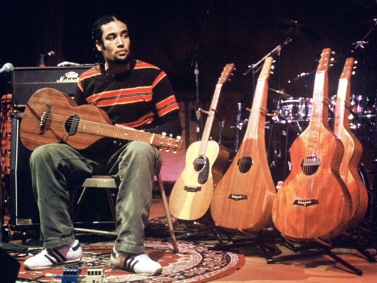 Ben Harper sits with a small guitar, surrounded by multiple acoustic guitars and drum equipment on stage.