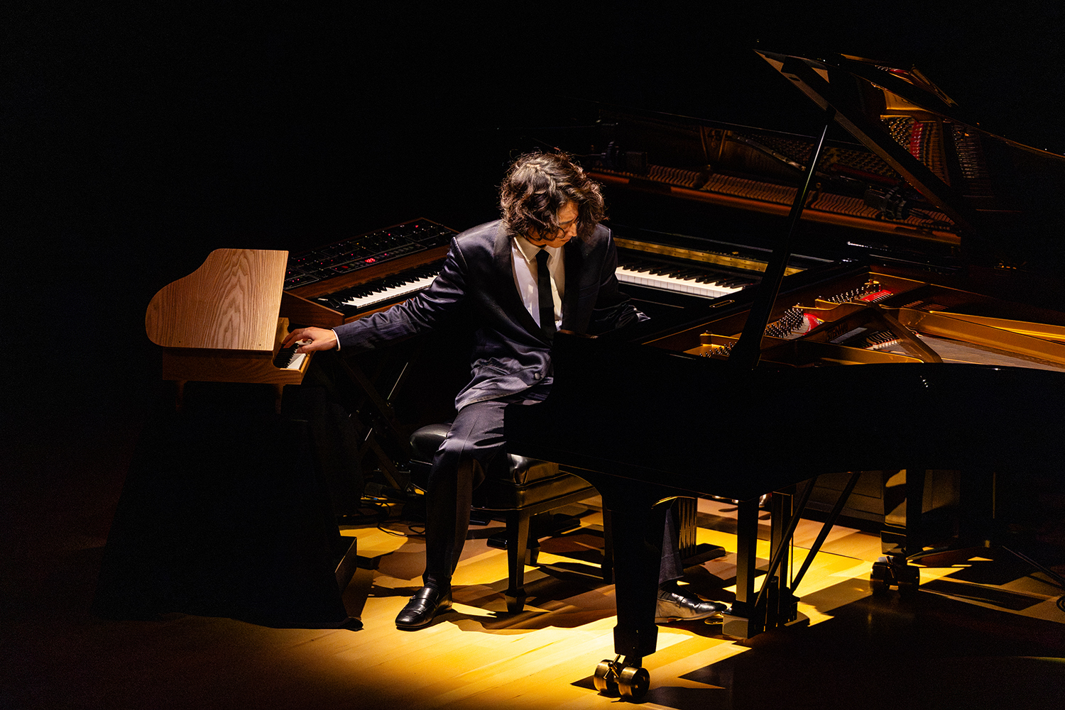 Hayato Sumino in a suit plays a grand piano and an electric keyboard under dramatic lighting on stage.