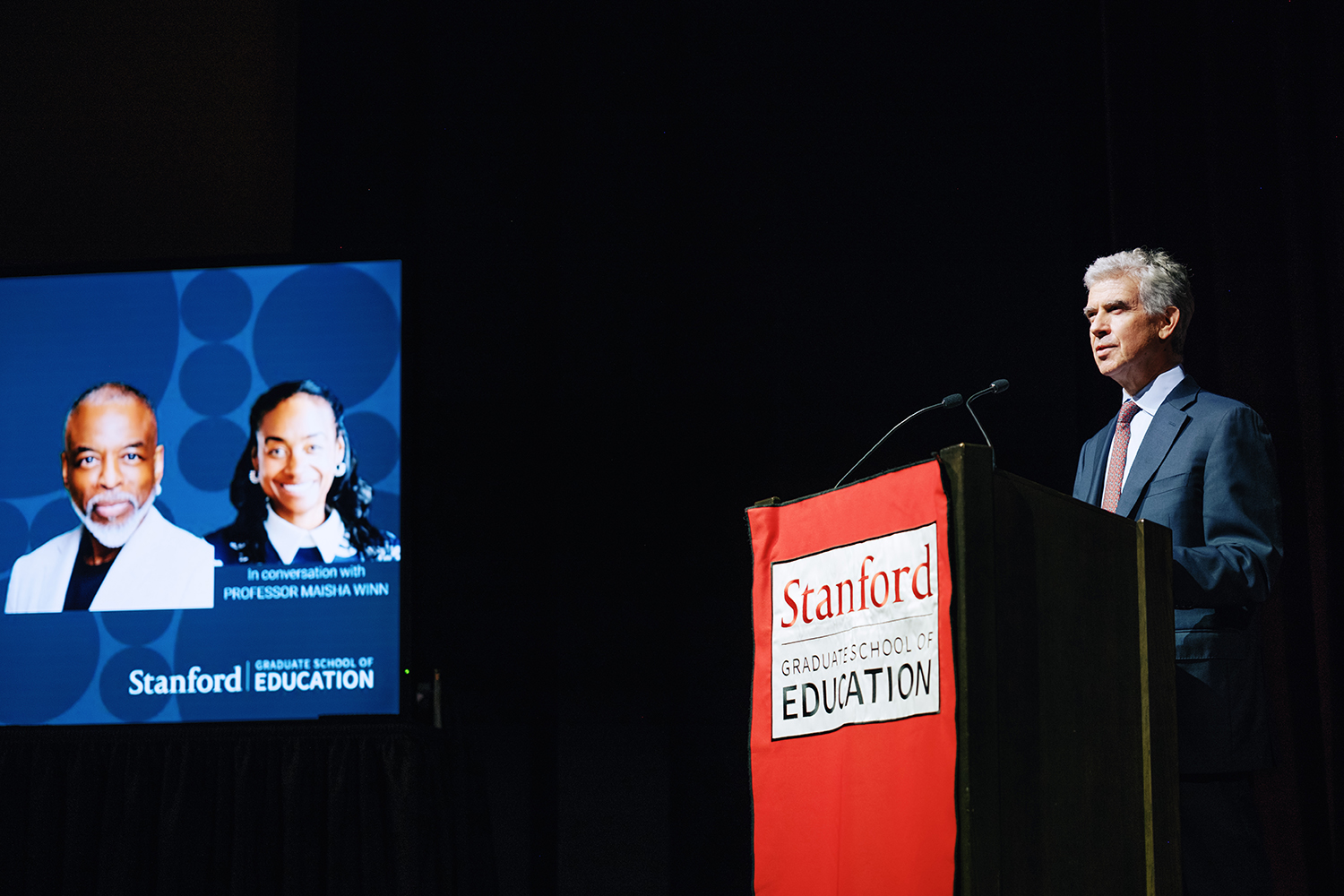 Dan Schwartz stands at a podium displaying the Stanford Graduate School of Education logo, while a presentation screen shows two people.