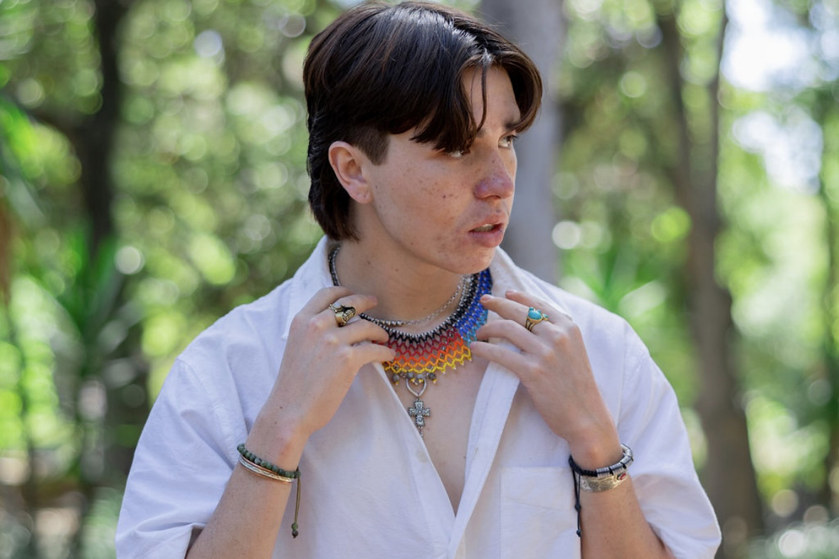 Ducan adjusting a colorful beaded necklace while standing outdoors with greenery in background.