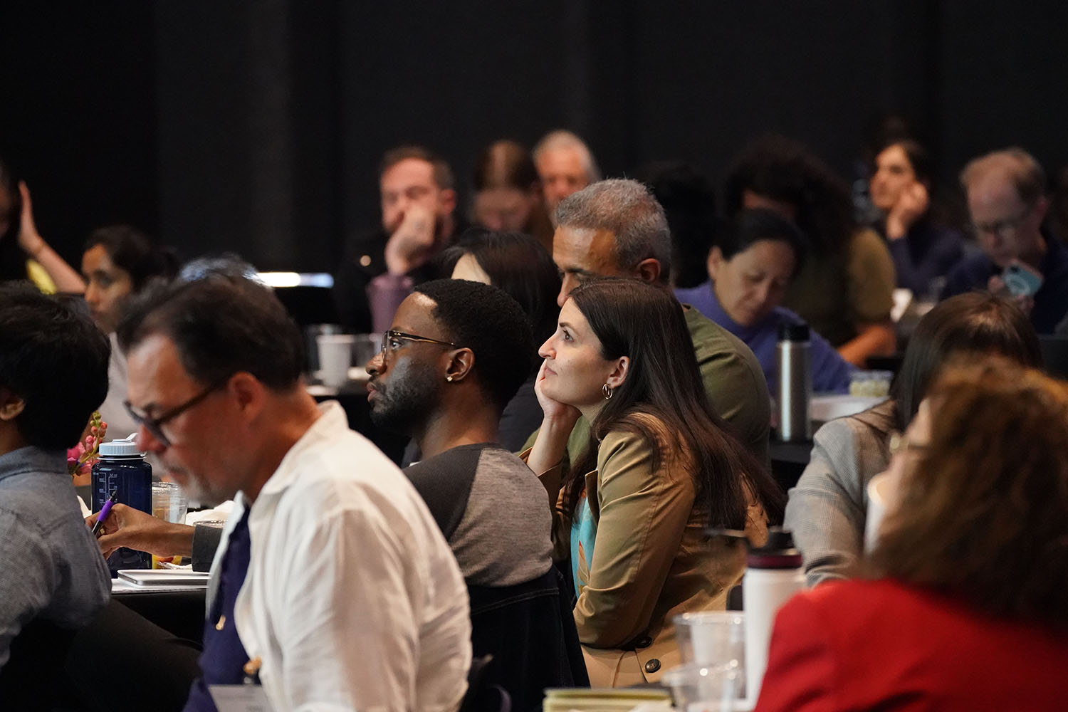 A diverse group of attendees listens intently at CORES Symposium, seated at tables with various items in front of them.