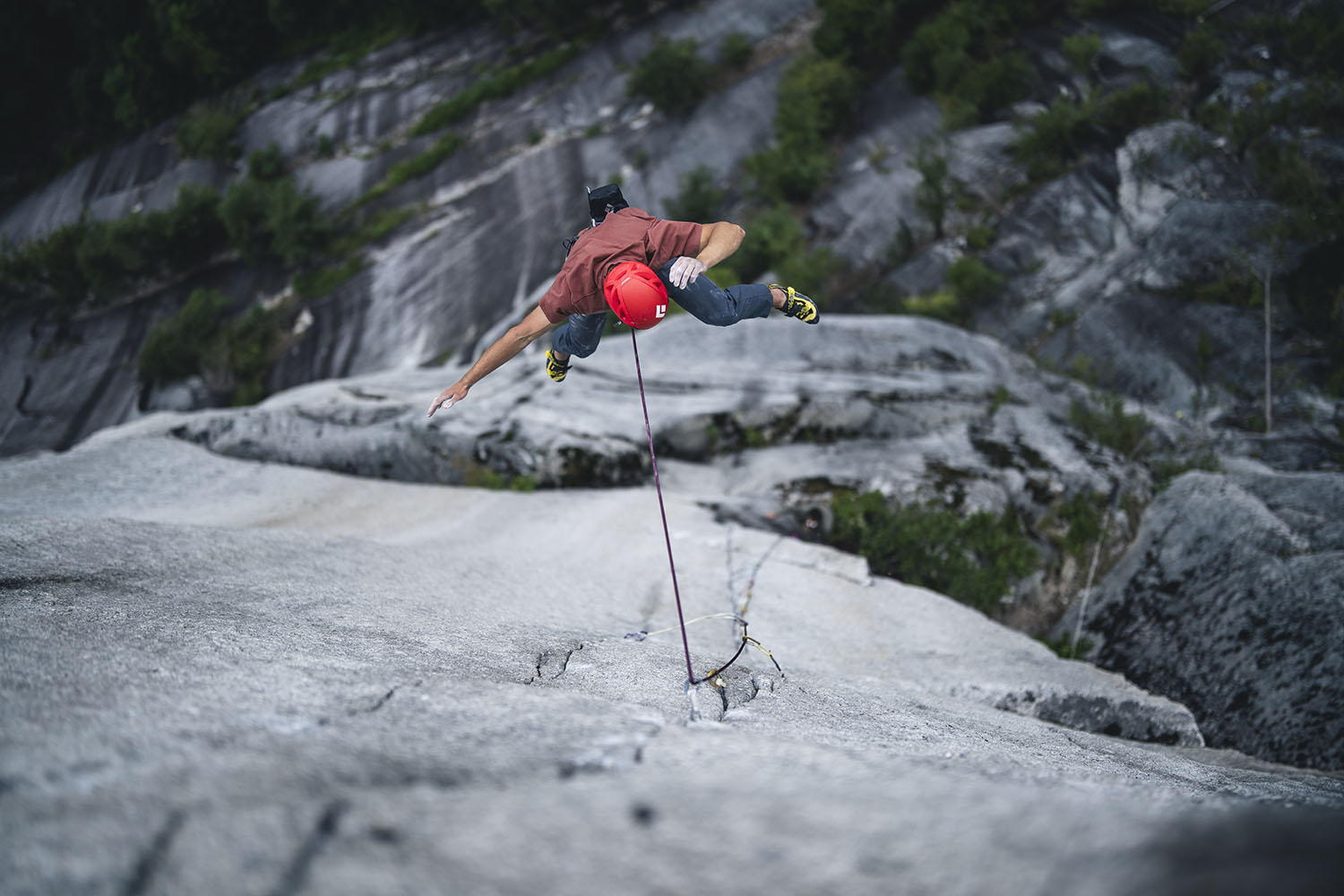 Climber Connor Herson in a red helmet leaps off a rock face, secured by a rope, against a background of rugged terrain and greenery.