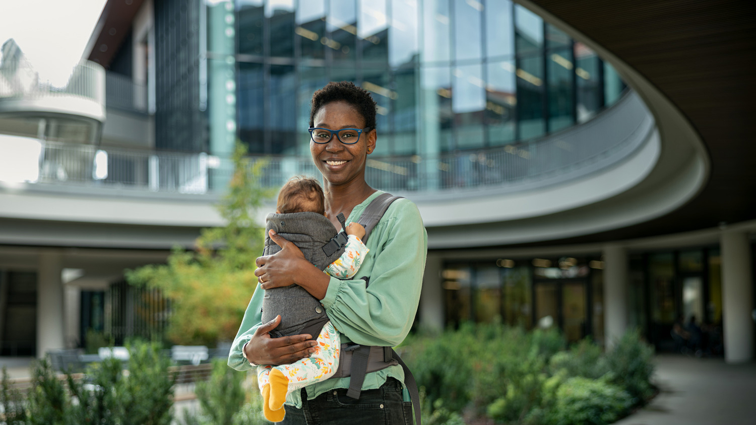 Hawa Racine Thiam holds an infant in a baby carrier while standing outside a modern building with large windows and greenery around.