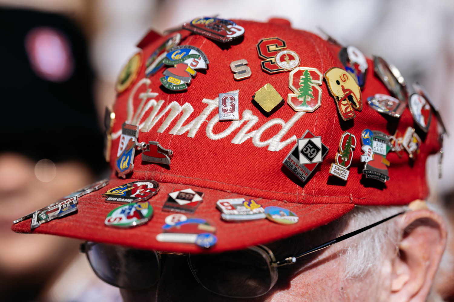 A red Stanford cap adorned with various colorful pins and badges representing different schools and events.