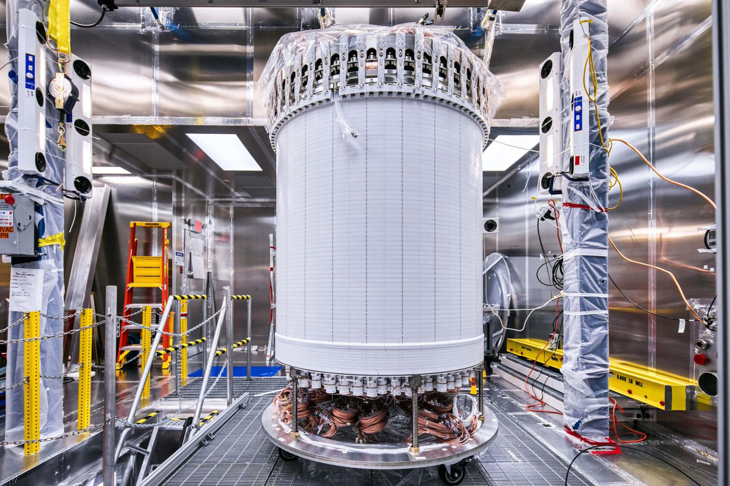 A tall white cylindrical particle detector wrapped in plastic sits on a cart inside a steel-lined clean room.