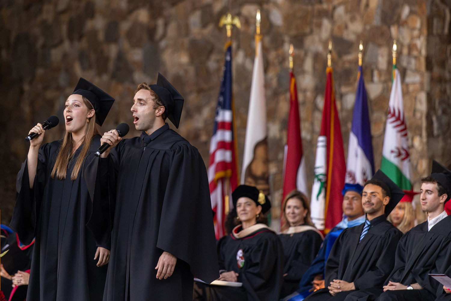 Two students in caps and gowns sing into microphones on stage, with ceremonial flags behind them.