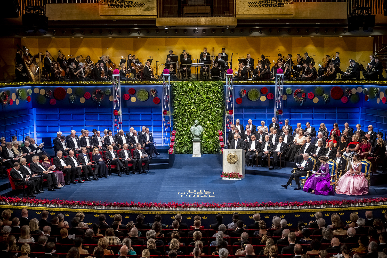 Wide view of the Nobel Prize ceremony stage, with laureates, dignitaries, an orchestra and a bust before a green wall.