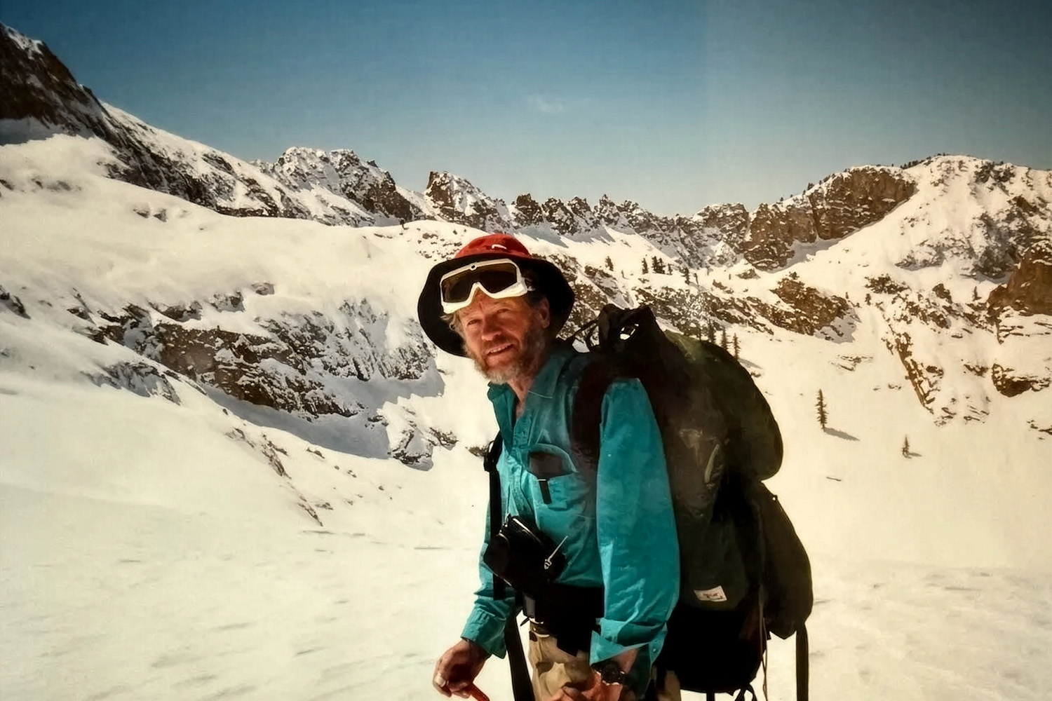 Peter Ray carries a large backpack across a snow-covered alpine basin ringed by rocky peaks.