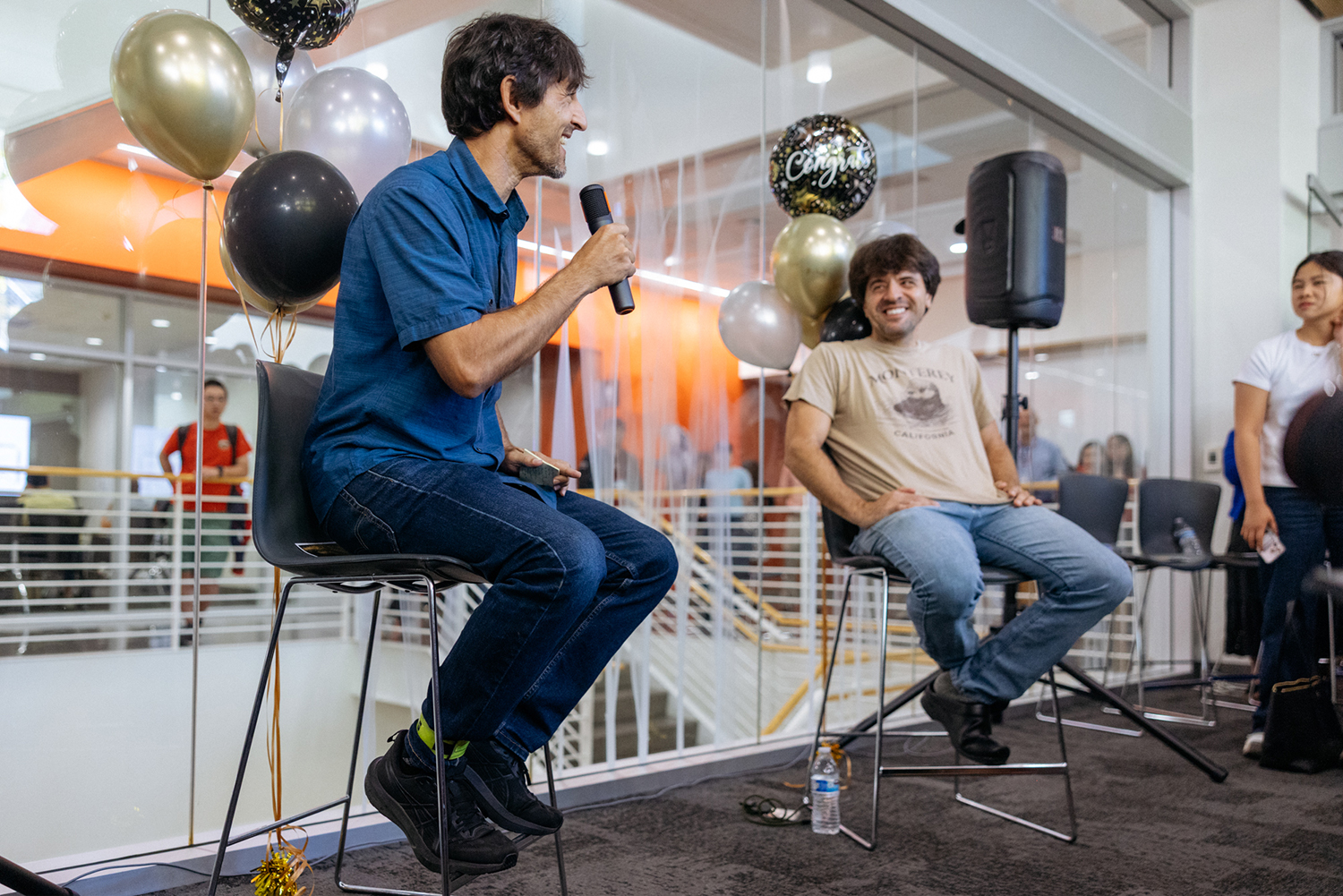 Two men sit on tall stools at a celebration, one speaking into a microphone and the other laughing, with gold, black, and silver balloons behind them.
