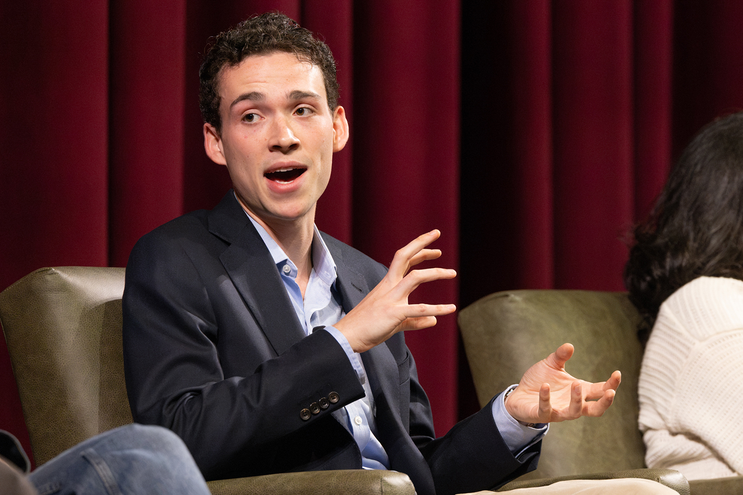 George Porteous speaks and gestures with both hands while seated on stage in front of a red curtain.
