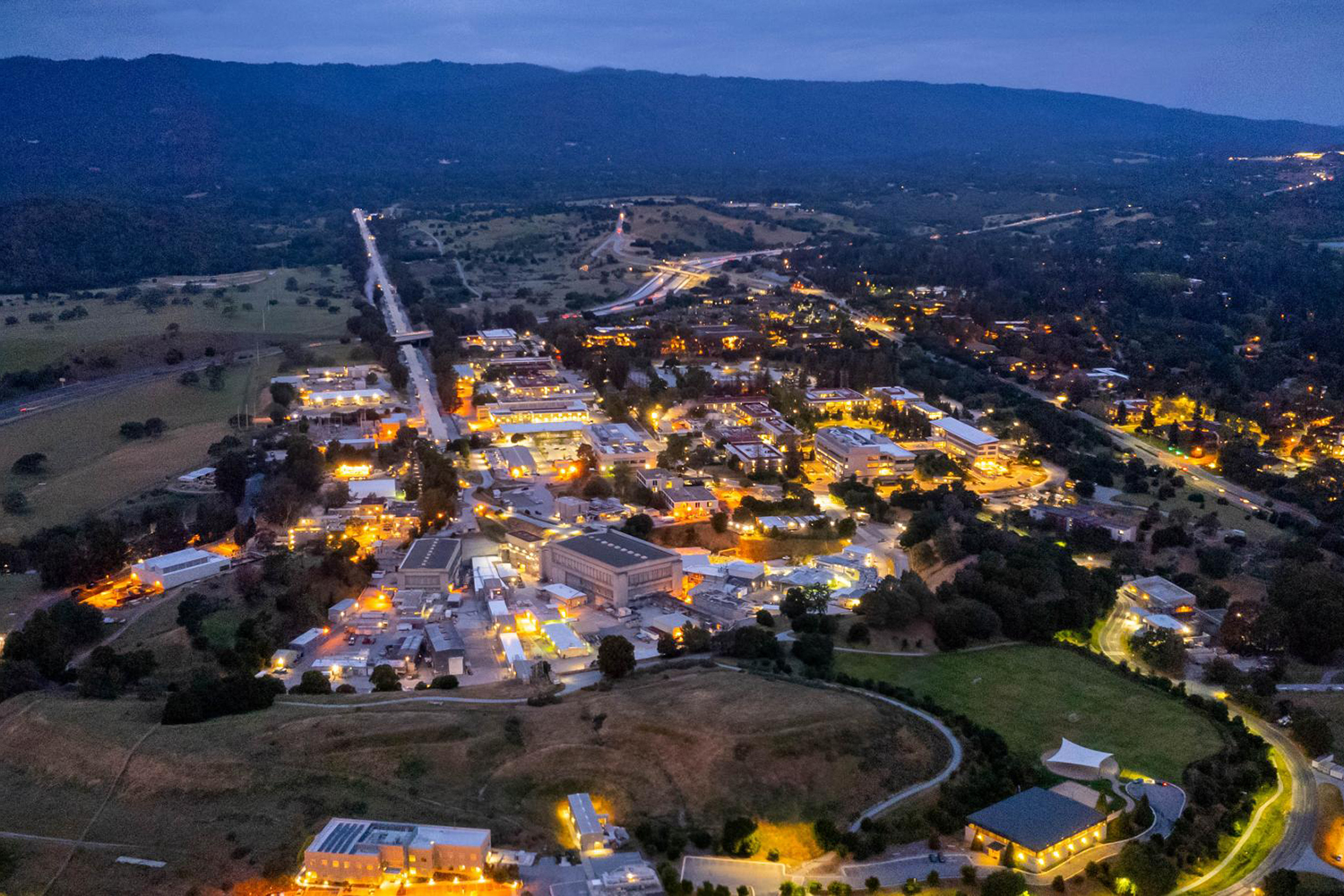 Aerial view at dusk of SLAC's facilities, with hills and a highway beyond.
