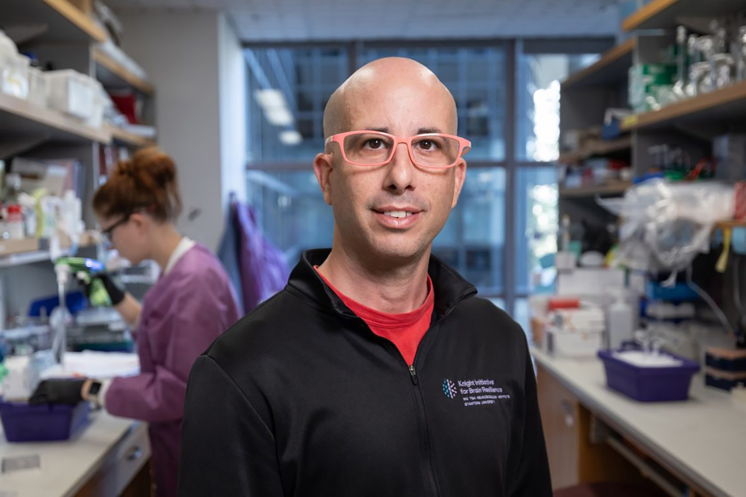 Aaron Gitler stands in a lab setting and poses for a photo, with a colleague working at a lab bench in the background.