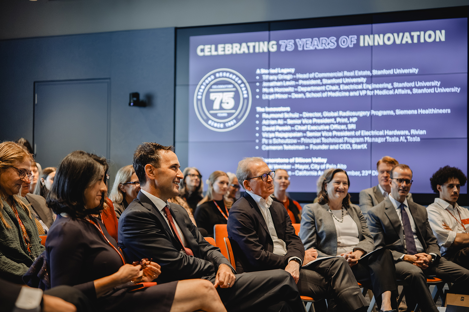 Audience members smile in front of a screen reading “Celebrating 75 Years of Innovation” and listing event speakers.