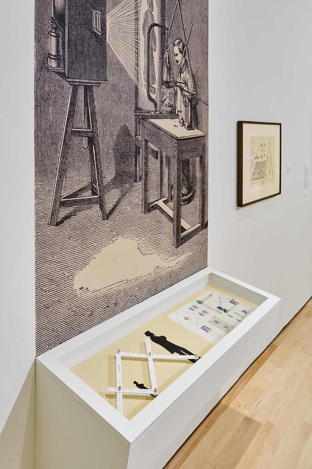 Antique illustration of an early photography studio installed on a gallery wall above a case of mechanical drawing tools.