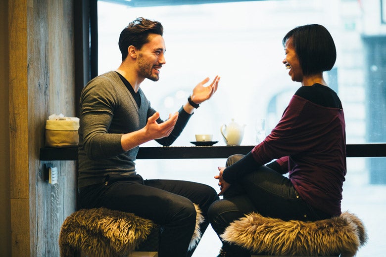 man with animated face making hand gestures while sitting across from a woman who is laughing