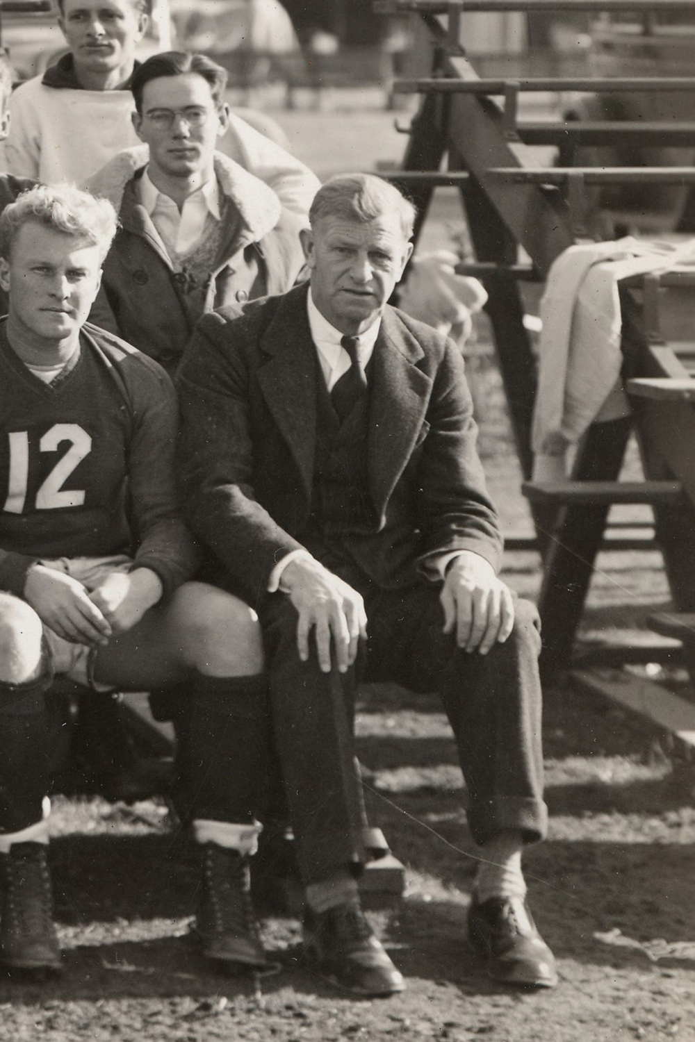 Pictured is a black and white photo of Harry Malone sitting on the edge of a bench next to a member of the Stanford rugby team. 